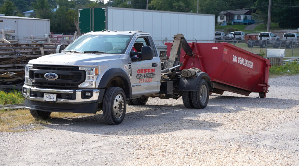 A silver Ford Super Duty truck with Griffin Waste Services branding is parked on a gravel area, towing a red dumpster rental container. Industrial buildings and a green hill are in the background.